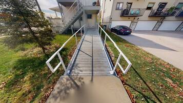A modern apartment building with a central staircase, a car parked in the driveway, and a tree to the left.