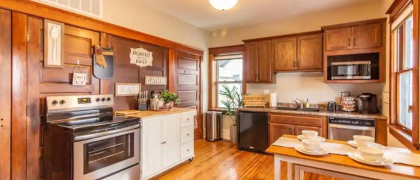 Una cocina con armarios de madera, una estufa y un microondas. Hay letreros en la pared y una mesa dispuesta con vajilla.