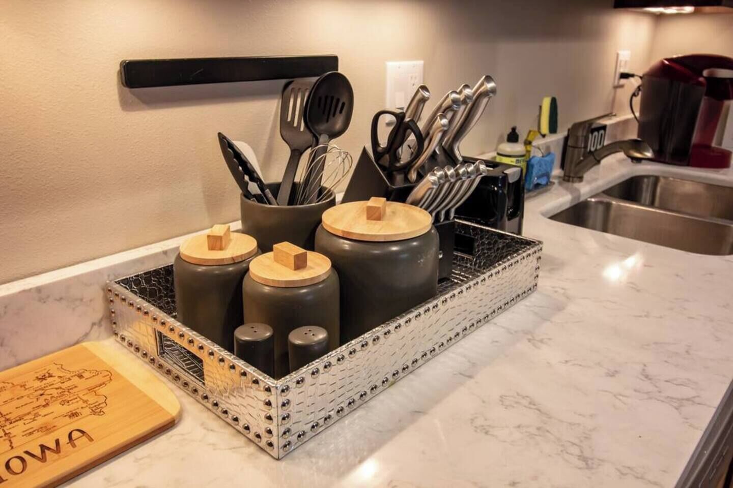 Una encimera de cocina con un especiero que contiene recipientes negros con tapas de madera, una tabla de cortar con texto y varios utensilios de cocina.