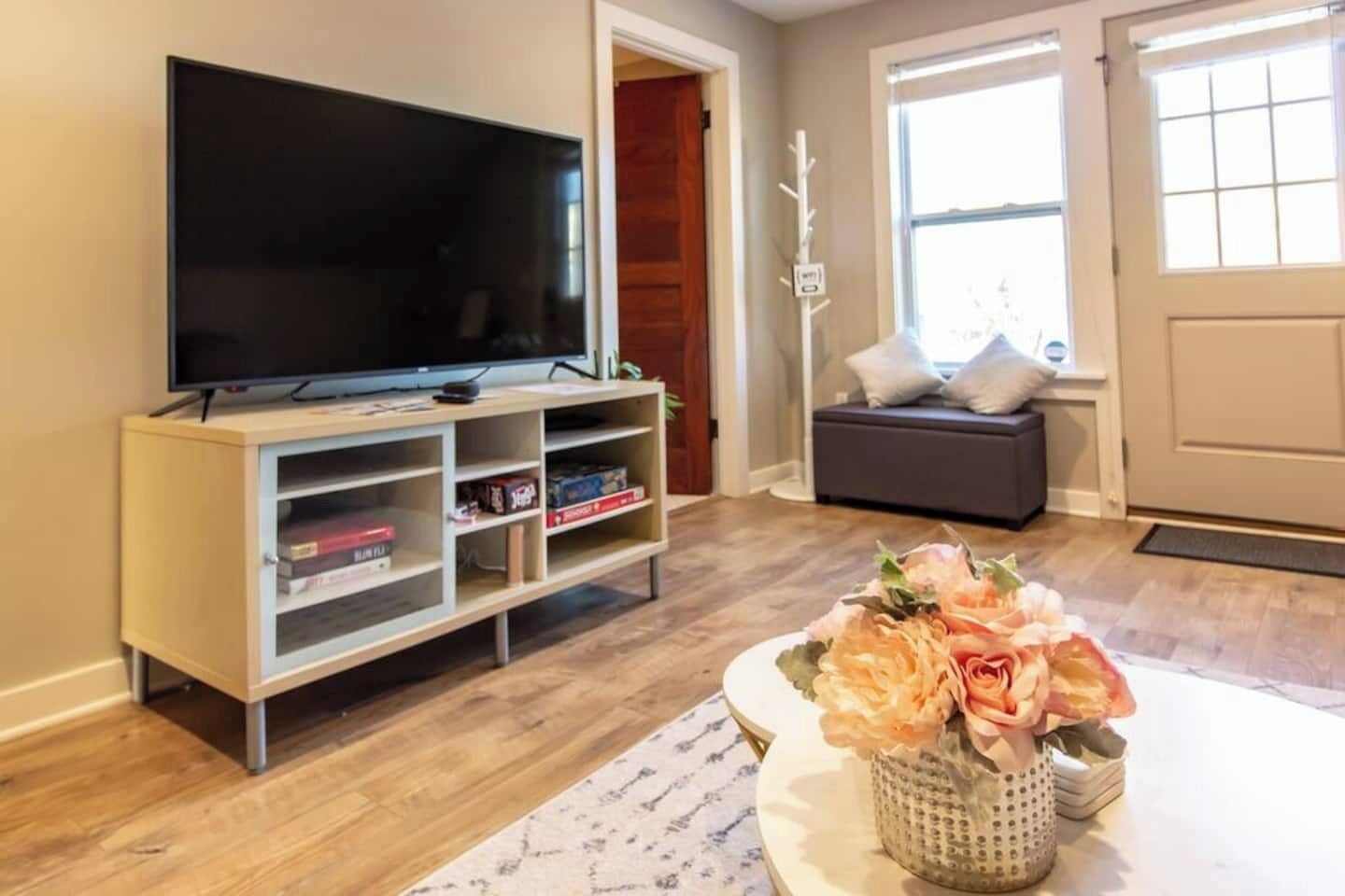 Ein Wohnzimmer mit Flachbildfernseher auf einem Schrank, einem Bücherregal, einem Esstisch mit Blumen in einer Vase und einem Fenster mit Blick nach draußen.