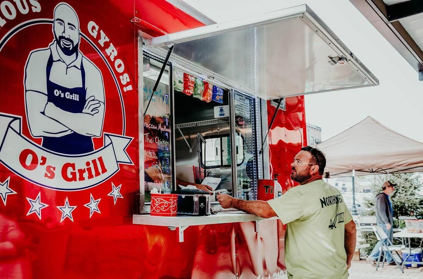 A food truck with a red and white logo, a man serving food, and a menu board visible.