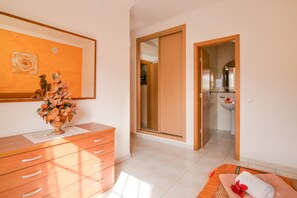 A hotel room with a wooden dresser, a vase of flowers, a painting, a sliding door to the bathroom, and a bed with a towel.