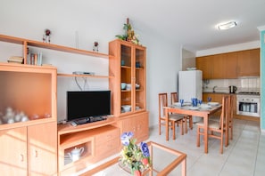 A modern kitchen with wooden cabinets, a dining table, and chairs. There is a television on a wooden stand and a cabinet with glass doors.