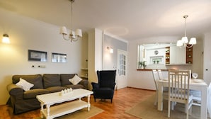 Un salon comprenant un canapé, un fauteuil, une table à manger et des chaises. On peut apercevoir la cuisine à travers une porte ouverte.
