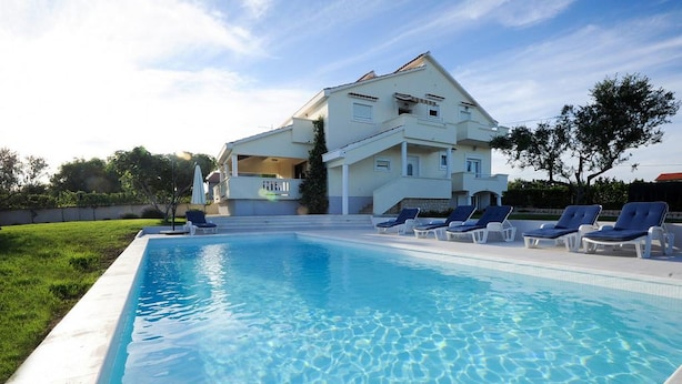 Une piscine avec des chaises longues, une maison en arrière-plan et un ciel dégagé.