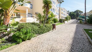 A paved pathway leading to a building with balconies, surrounded by greenery and palm trees.
