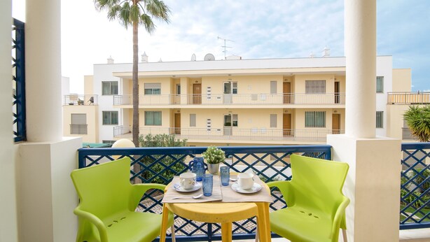 A balcony with two green chairs, a small table with a vase, and a view of a multi-story building.