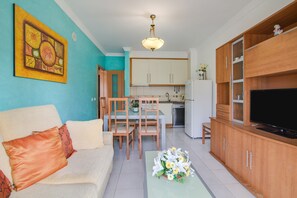 Una sala de estar con sofá, mesa de centro con flores, televisor, armarios de madera, comedor con sillas y cocina con alacenas y lavaplatos.