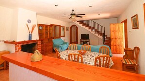 Un comedor con una mesa y sillas de madera, una cocina con estufa de ladrillo y una escalera que sube a otro nivel.