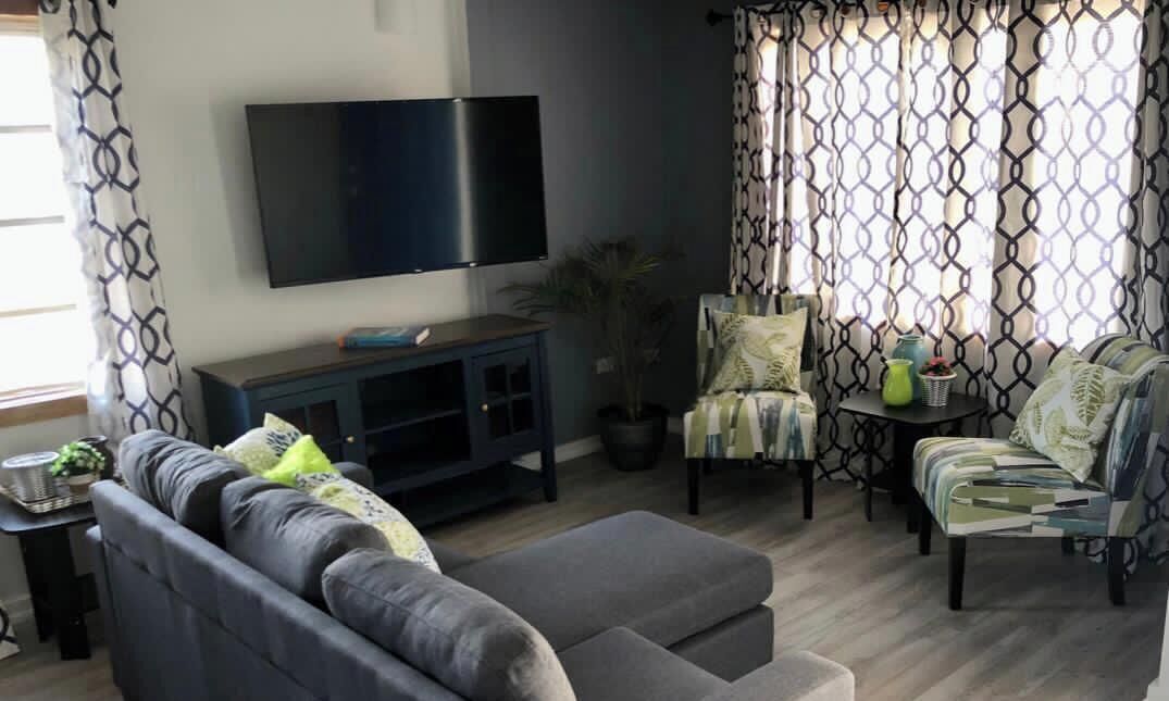 Ein Wohnzimmer mit Sofa, Sessel, Hocker, Fernseher und gemusterten Vorhängen.