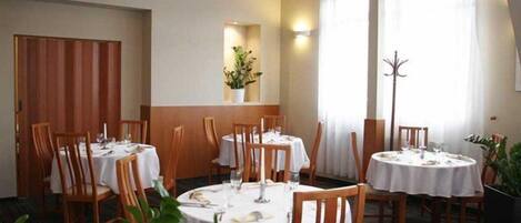 A restaurant with round tables and wooden chairs, a potted plant, and a window with curtains.