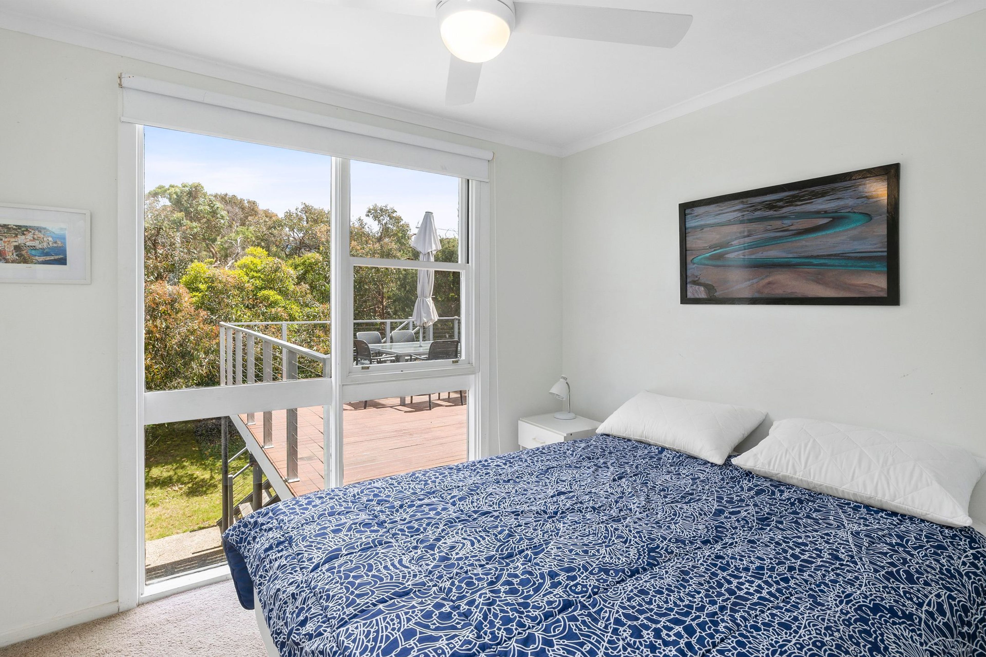 A bedroom with a large bed, a nightstand, a desk, and a window overlooking a deck.
