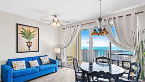 A living room with a blue sofa, a dining table with chairs, a ceiling fan, and a large window with curtains. - ☀️Pools-18th Fl Gulf Views-1BR+Bunks Ariel Dunes II 1805☀️ (Destin)