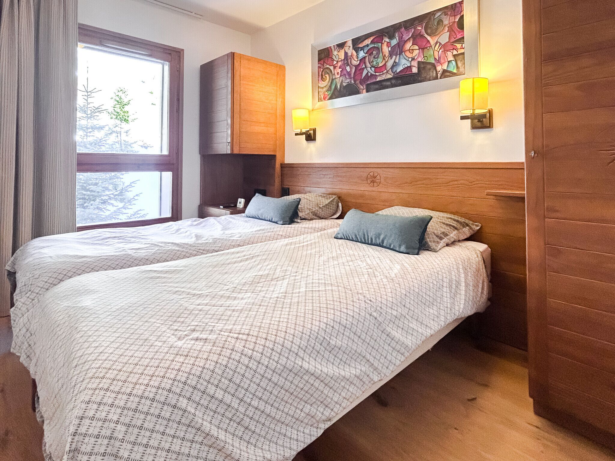 A double bed with a patterned bedspread, a nightstand with a lamp, a window with curtains, and a wooden headboard.