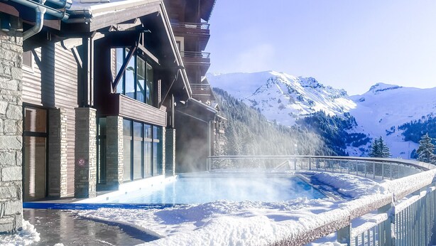 Un'area benessere con piscina, circondata da montagne innevate e un edificio in legno con ampie finestre.