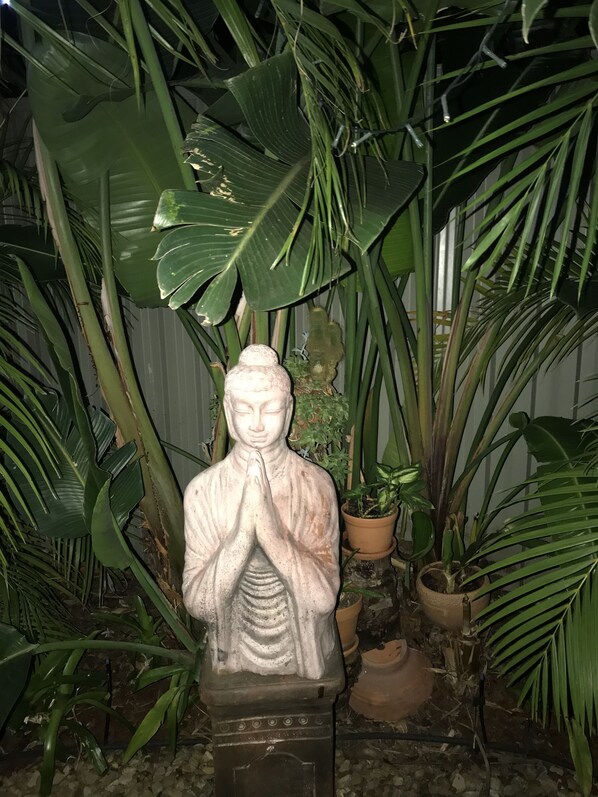 A statue of a figure in a prayer pose, surrounded by large green leaves and potted plants.