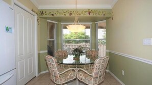 Een eetgedeelte met een glazen tafel, omringd door stoelen met een bloemenprint, een hanglamp en uitzicht door een raam met jaloezieën.