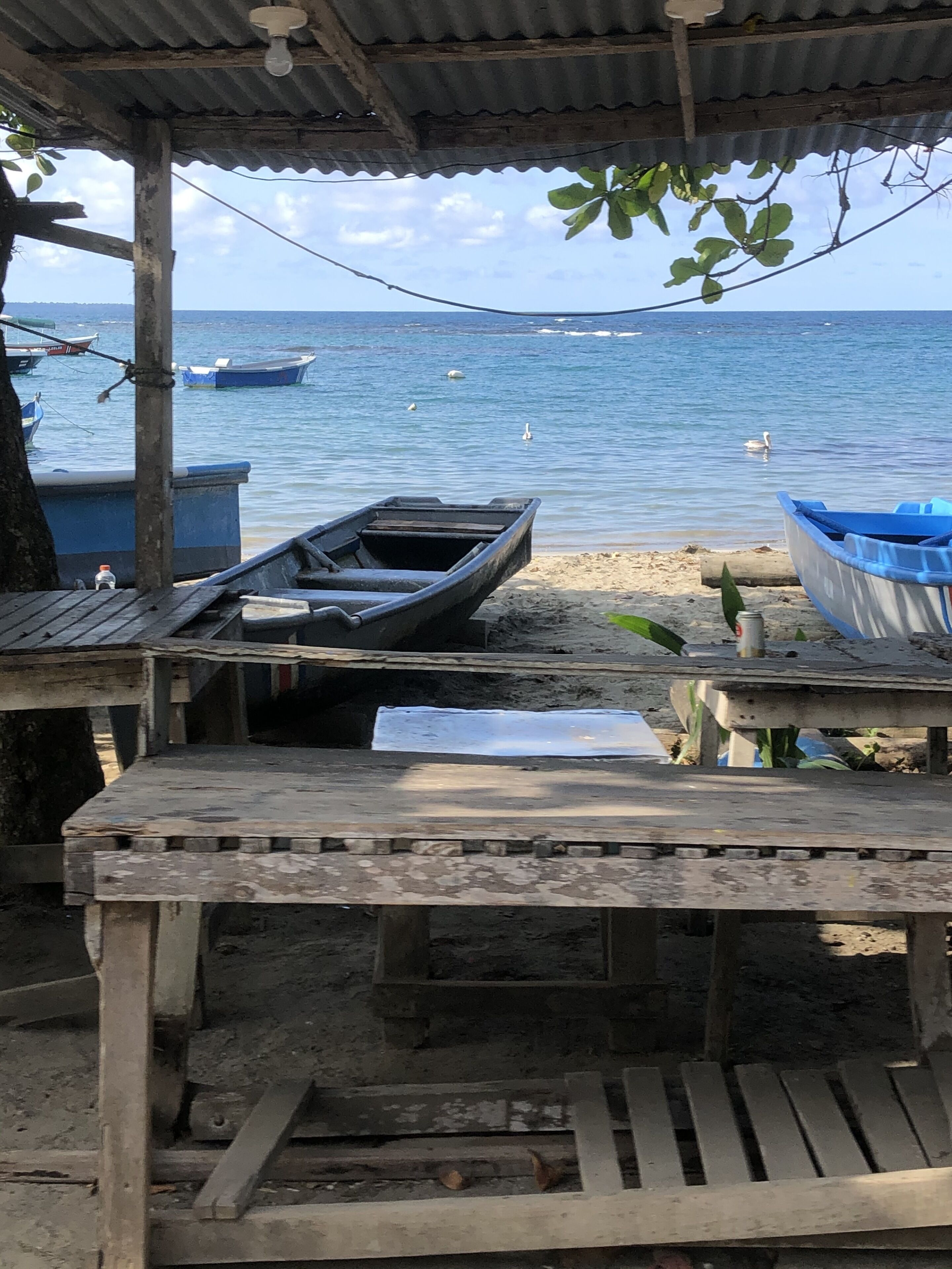 Ein rustikaler Holztisch mit Bänken unter einem Wellblechdach mit Blick auf ein ruhiges Meer, an dessen Ufer mehrere Boote vor Anker liegen.