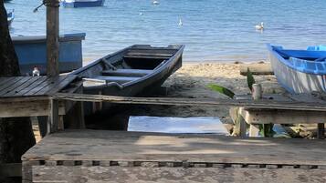 Ein rustikaler Holztisch mit Bänken unter einem Wellblechdach mit Blick auf ein ruhiges Meer, an dessen Ufer mehrere Boote vor Anker liegen.