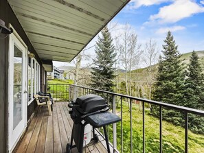 Un terrazzo in legno con barbecue, sedie e vista su alberi e colline.