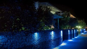 A stone wall with blue lighting along a pathway leading to a building with a sloped roof. - Luxury Three Bedroom apartment with hot tub "L&E" (Santorini)