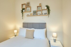 A neatly made bed with a grey headboard, two bedside lamps, and a wall-mounted shelf with framed pictures and plants.