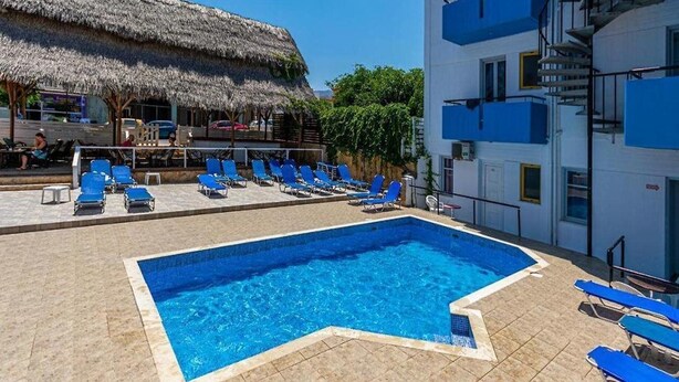 Zwembad met blauw water, omringd door blauwe ligbedden. Er is een constructie met rieten dak en een wit gebouw met blauwe balkons.