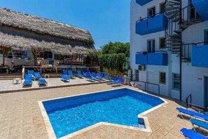 Una piscina con agua azul, rodeada de sillas reclinables azules. Se observa una estructura con techo de paja y un edificio blanco con balcones azules.