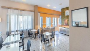 Een restaurantgedeelte in een hotel, met tafels en stoelen, een buffet en een groot raam met gordijnen.
