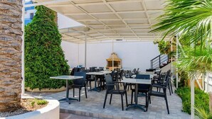 Een terras buiten met tafels, stoelen, een grote groene struik en een barbecue.