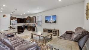 A modern living room with a brown leather sofa, a wooden coffee table, and a kitchen area with dark wooden cabinets and a white refrigerator. - Luxury 8BR Villa  Private Pool, Game Room ,Near Disney , Windsor Island Resort (Davenport)