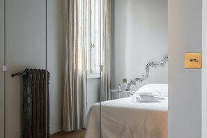 A hotel room with a bed, a radiator, a window with curtains, a bedside table, and a wall-mounted light switch. - Cà Bellavista (Venice)