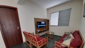 A living room with a wooden door, a television mounted on the wall, a window with blinds, and two wicker sofas with red cushions.