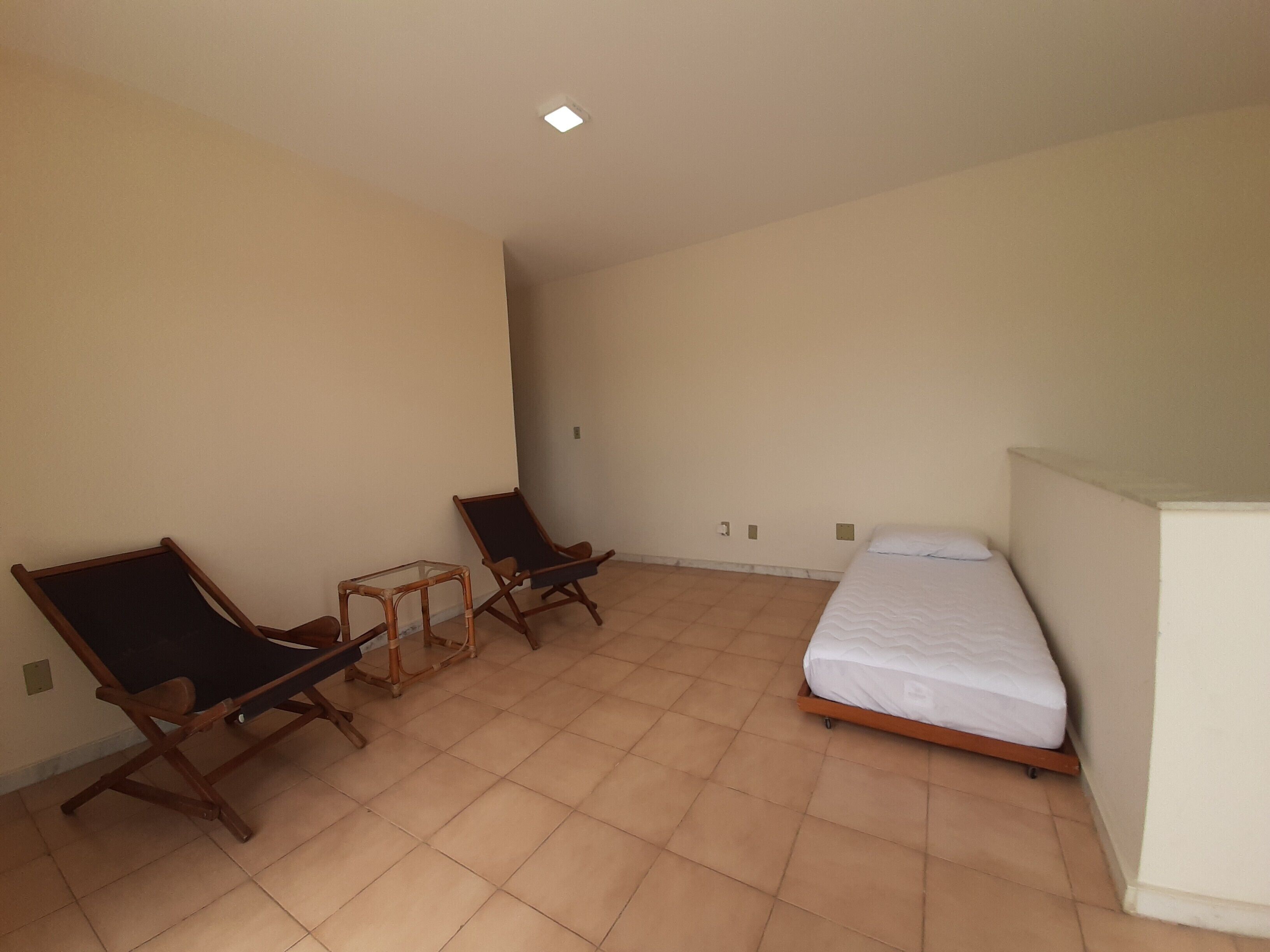 Quarto pequeno e pouco mobiliado, com uma cama, duas cadeiras de madeira e uma mesa.