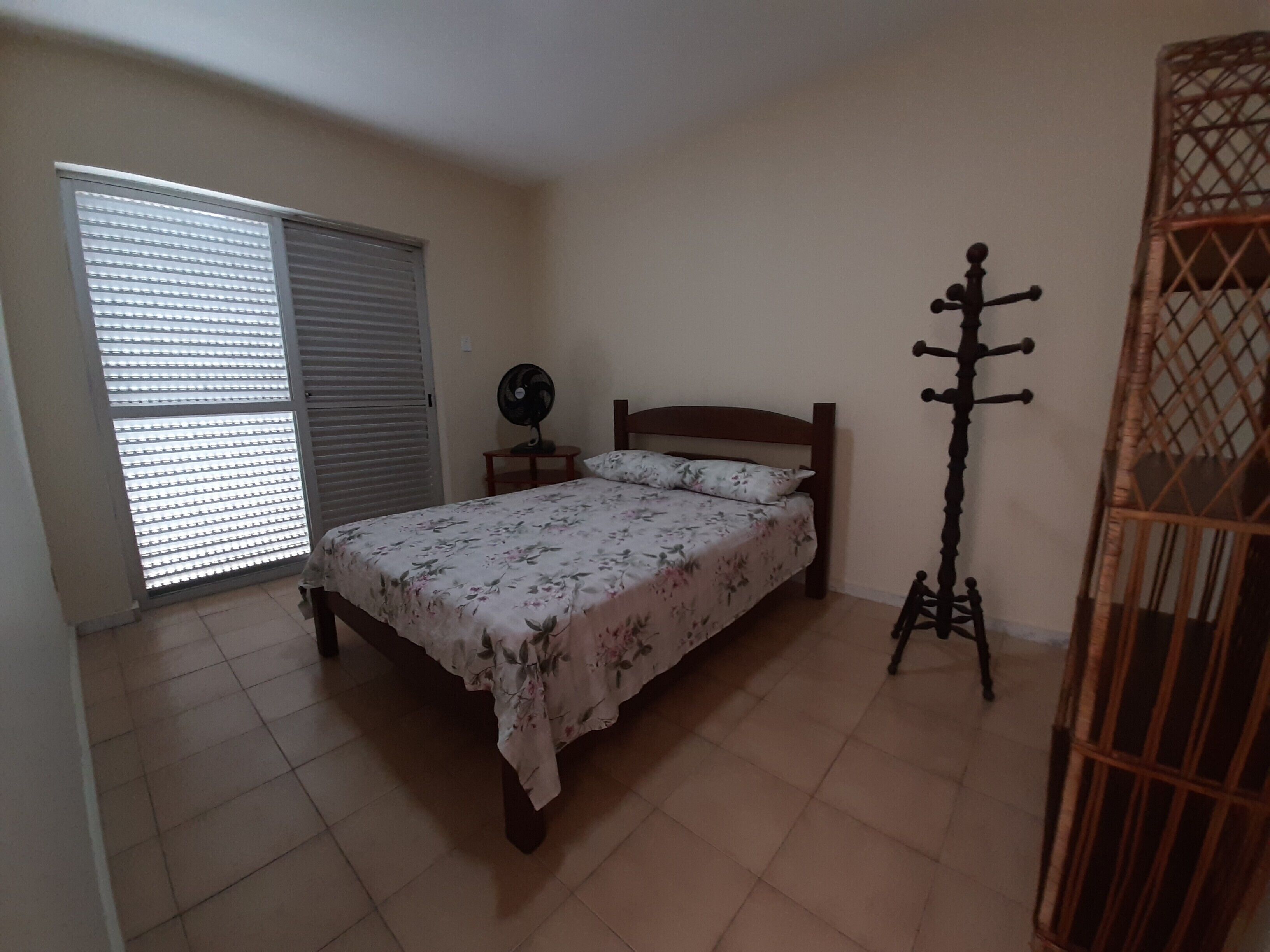 A bedroom with a bed, a standing fan, a window with blinds, and a wicker chair.