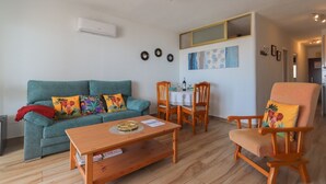 Ein Wohnzimmer mit einer Couch, einem Couchtisch und einem Esstisch mit Stühlen. Es gibt einen Deckenventilator, eine Klimaanlage und Wanddekorationen.