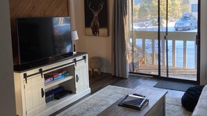 A living room with a TV, a wooden wall panel, a deer painting, and a view of a snowy landscape through a sliding glass door. - Cozy Condo in a great location! (Mammoth Lakes)
