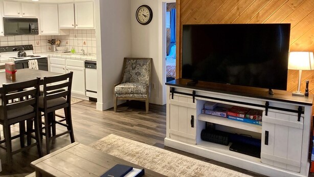 A modern living room with a TV, a dining area with a table and chairs, a patterned armchair, and a kitchen with white cabinets and a microwave. - Cozy Condo in a great location! (Mammoth Lakes)