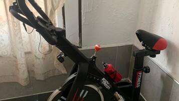 Ein stehendes Ergometer steht in einer Ecke eines Raumes mit Fliesenboden und einem Vorhang im Hintergrund.