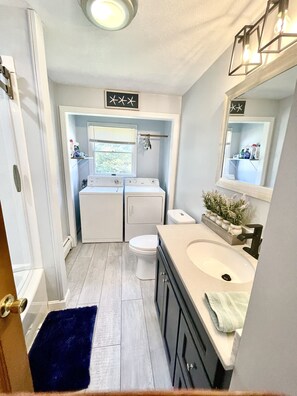 A compact bathroom with a white toilet, a double washer-dryer unit, a sink with a mirror, and a blue rug. - Bonnet Shores 6 Bed  3 Bath in Bonnet near weekend Neighborhood shuttle to Beach (Narragansett)