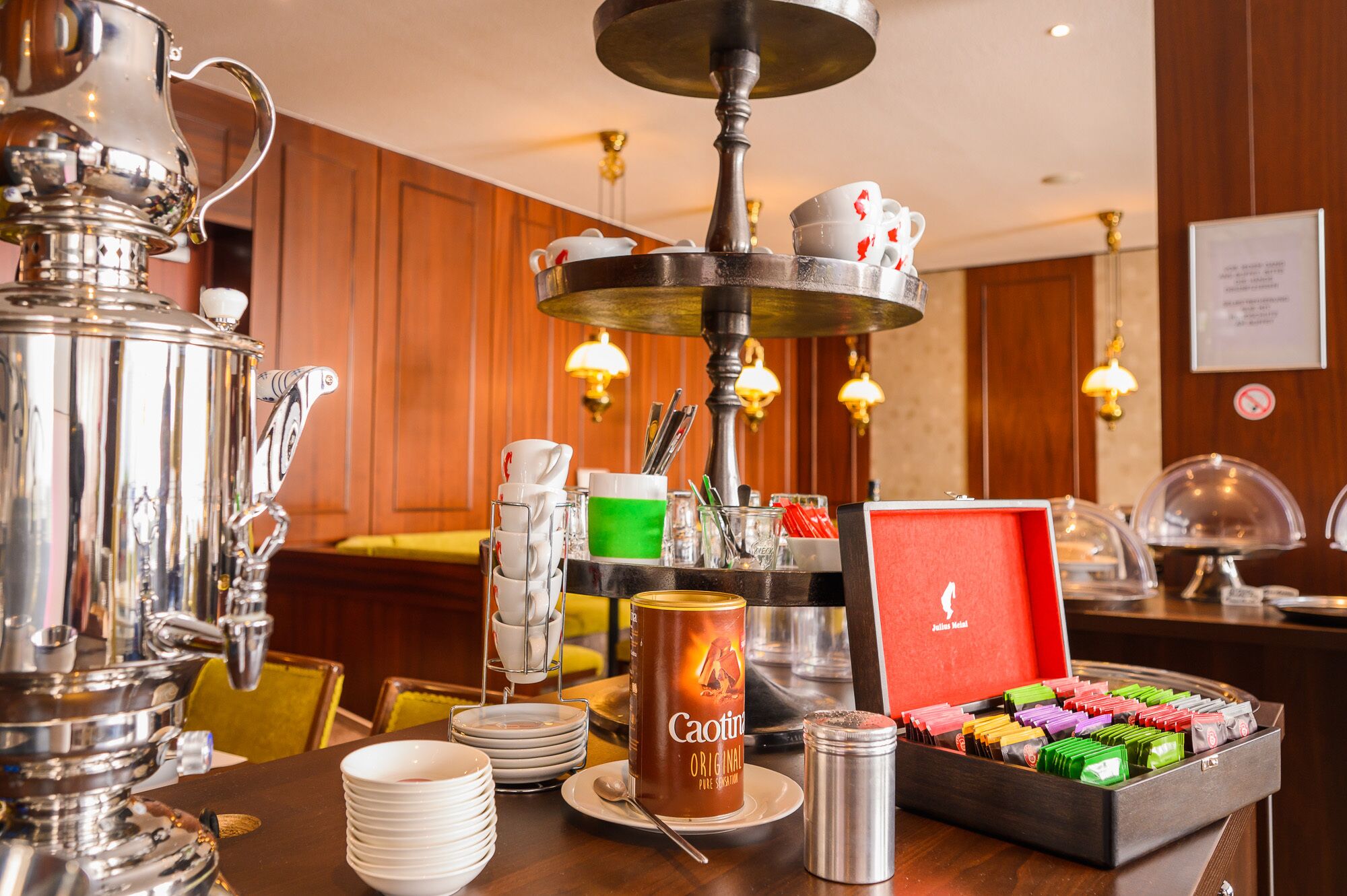 Una mesa de buffet con un dispensador de café, platos apilados, una lata de café y una caja de bombones variados.