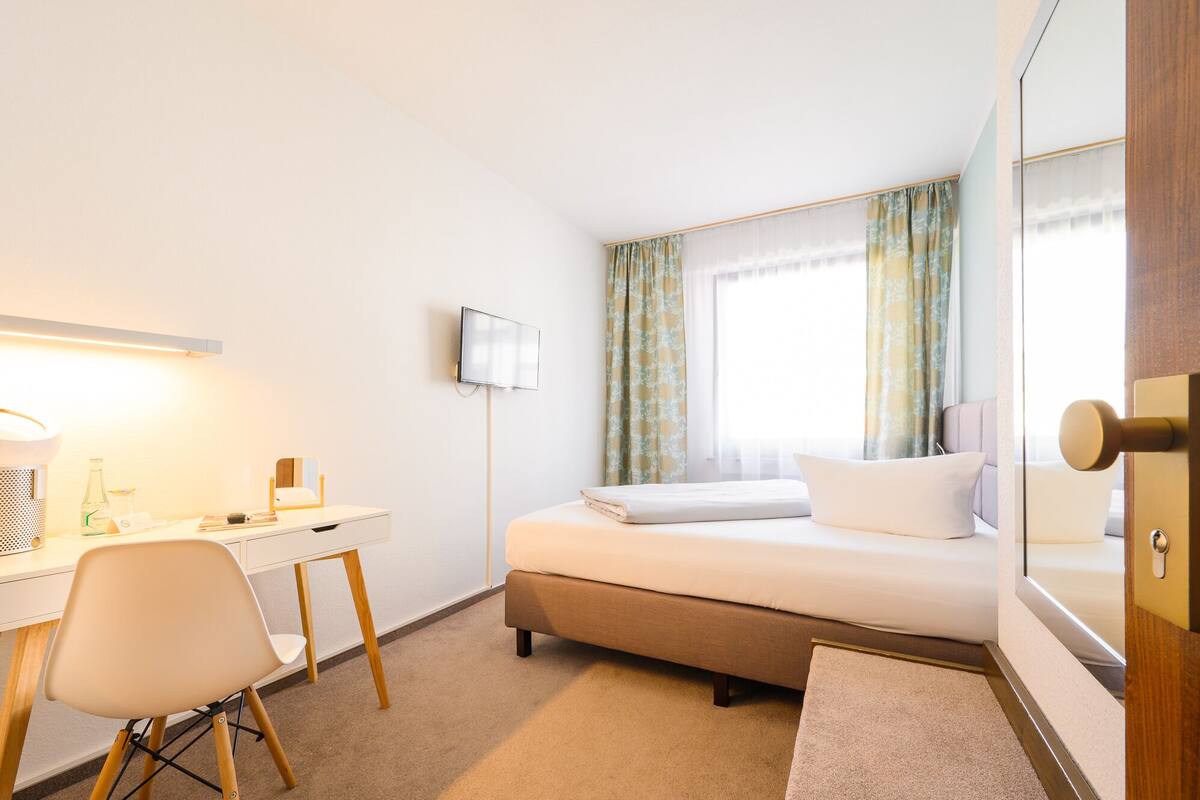 Une chambre d’hôtel avec un lit, un bureau et une chaise, une télévision et une fenêtre avec des rideaux.
