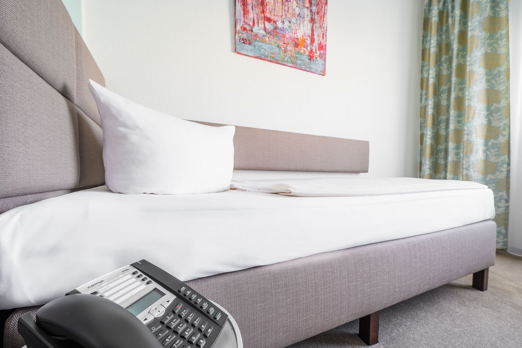 A modern bedroom with a grey sofa, white bedding, and a small bedside table with a telephone.
