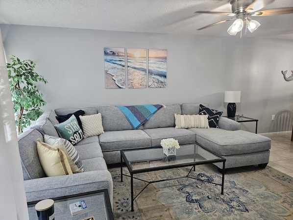 A modern living room with a grey sectional sofa, a glass coffee table, and a large painting of a beach scene. - Great Island Getaway! (Holmes Beach)