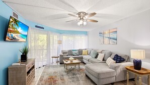 A living room with a ceiling fan, a sofa, a coffee table, and a TV on the wall. - Great Island Getaway! (Holmes Beach)