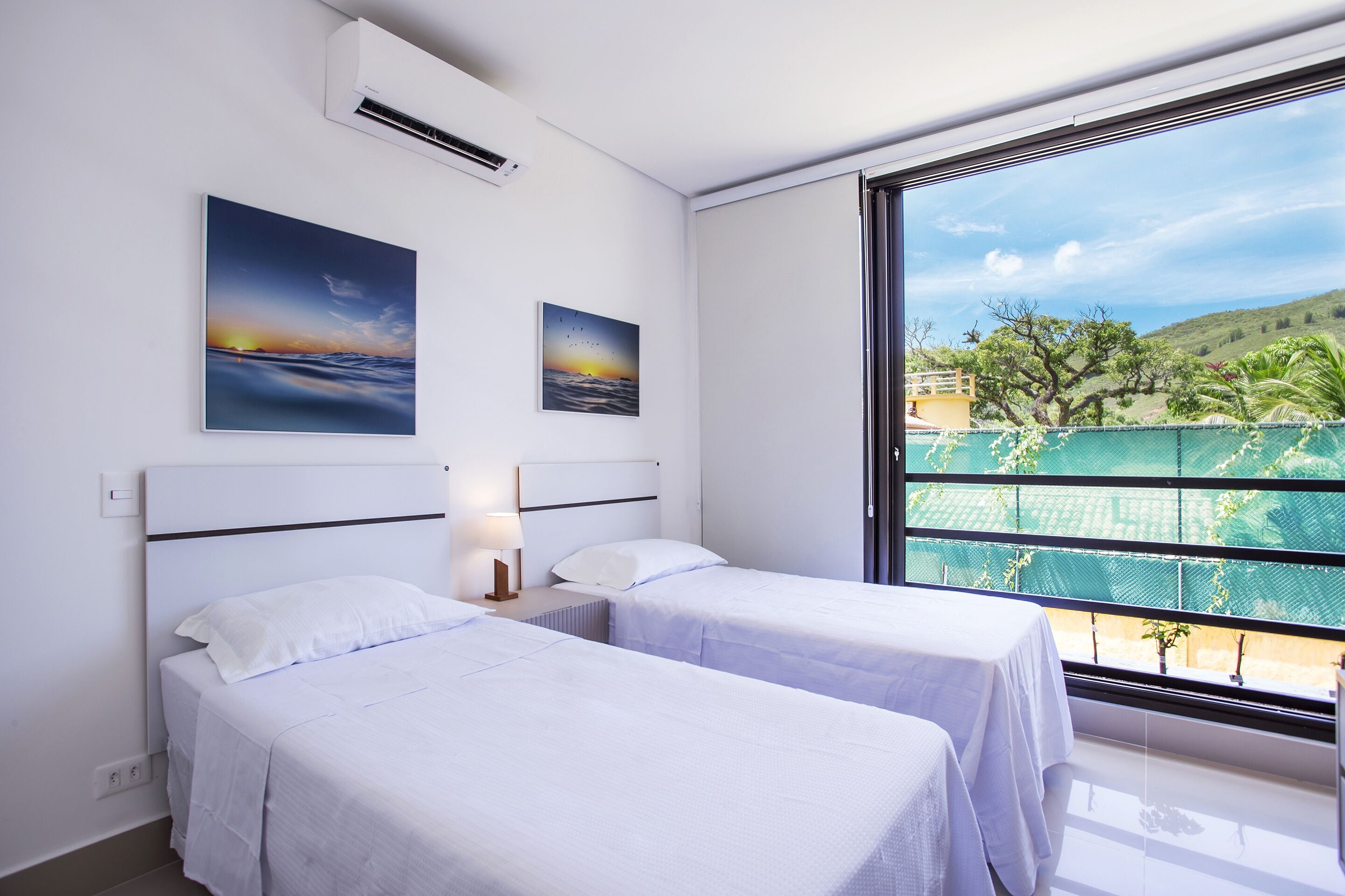 A hotel room with two beds, a large window with a view of a green landscape, and two paintings on the wall.