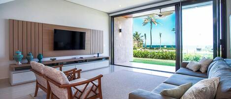 A modern living room with a flat-screen TV, wooden furniture, and a large sliding glass door leading to a balcony with a view of palm trees and the ocean.