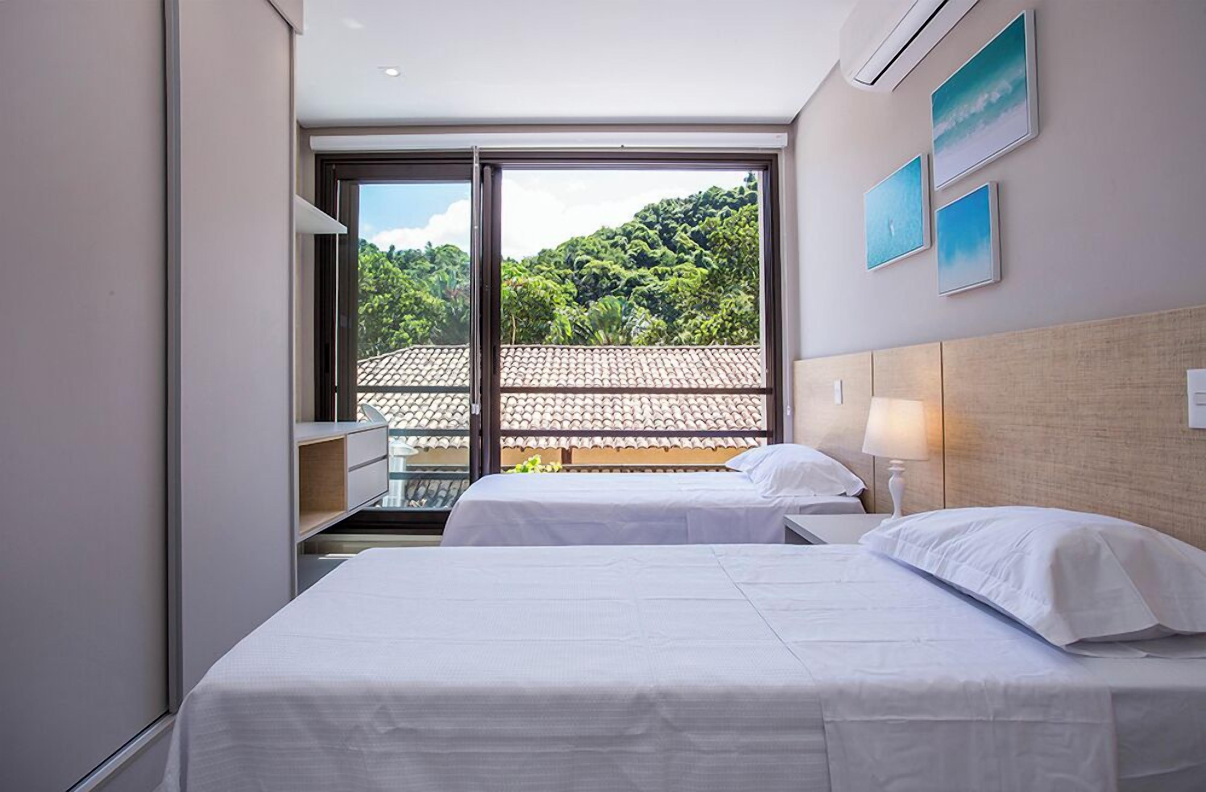A hotel room with two beds, a large window with a view of a tiled roof and greenery, and wall-mounted artwork.