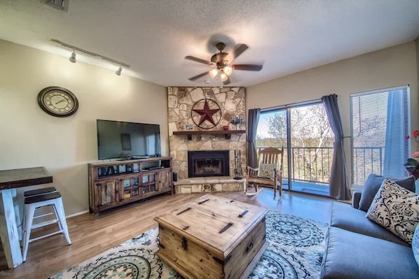 A living room with a stone fireplace, a television, a ceiling fan, and a large window. - SS B11 Seele Street River Retreat (New Braunfels)
