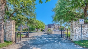 A gated entrance with stone pillars, a clear sky, and a parking area with cars. - SS B11 Seele Street River Retreat (New Braunfels)